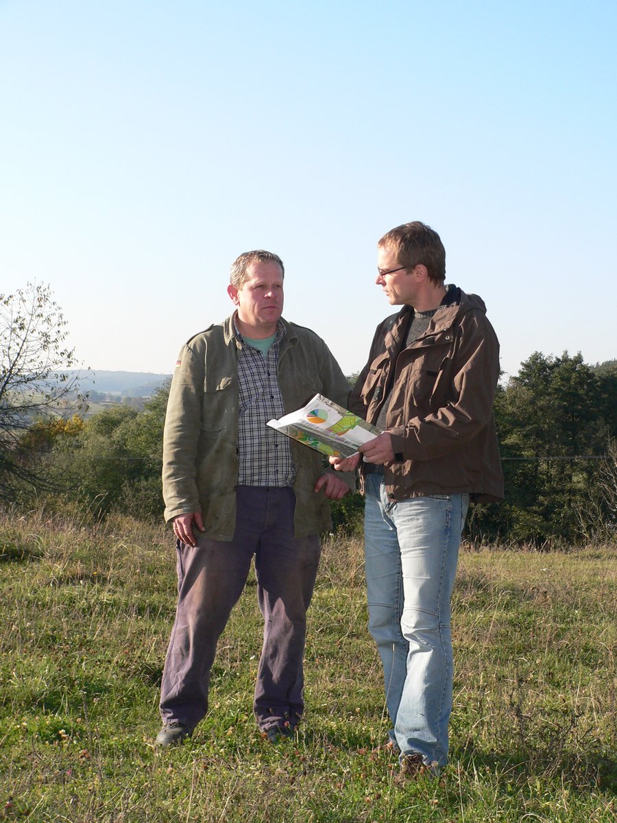 Zwei Männer stehen auf einer Wiese. Einer hält einen Karte in den Händen. Im Hintergrund eine Hecke. 