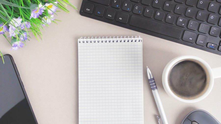 Tastatur mit Maus, Handy, Notizblock, Stift, Kaffeetasse und Blumen.